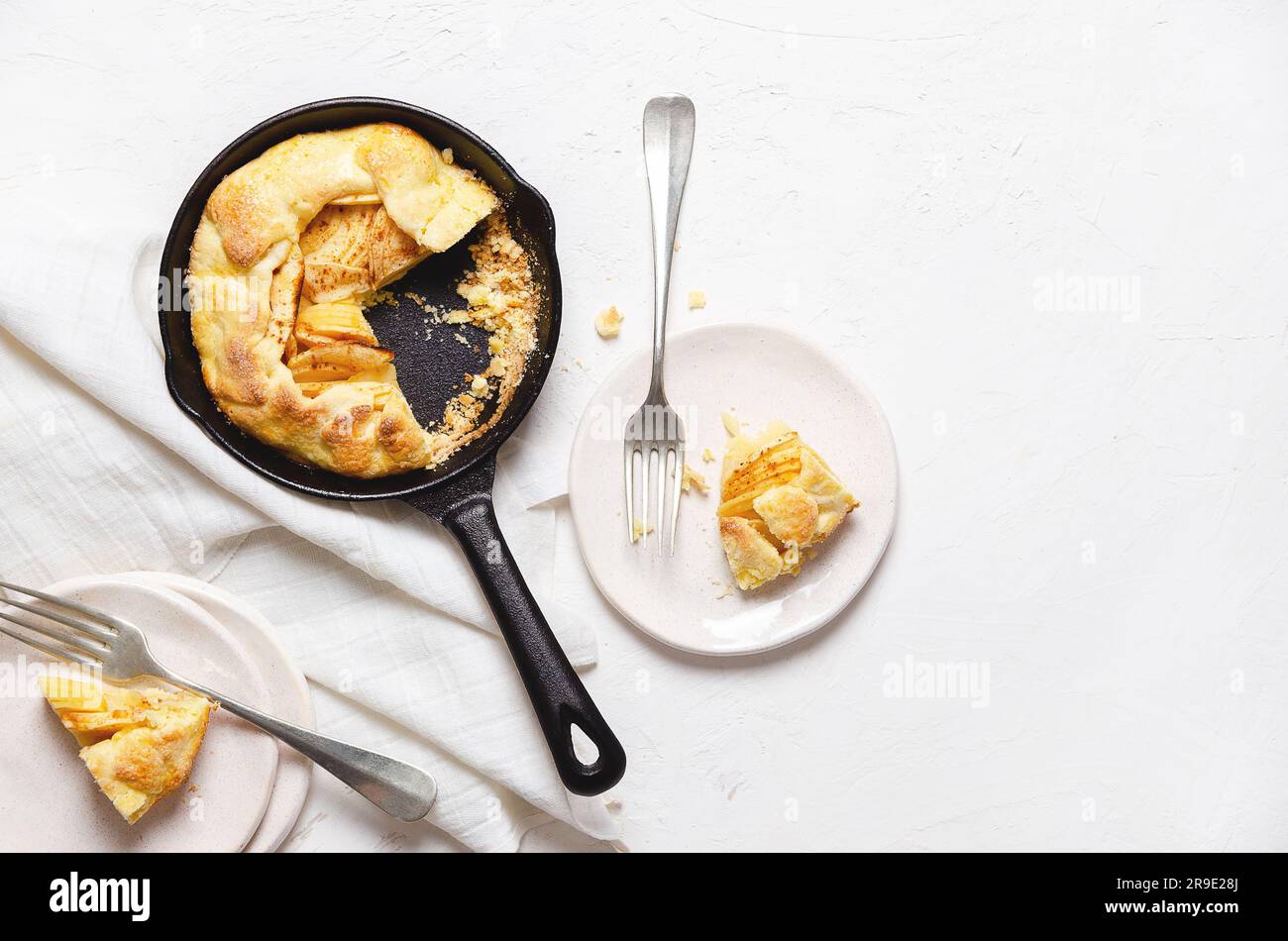 Apfelgalette in einer schwarzen Pfanne, zwei Galette in weißen Tellern mit Gabeln und ein Stück weißes Tuch auf weißem Hintergrund mit Kopierraum. Stockfoto