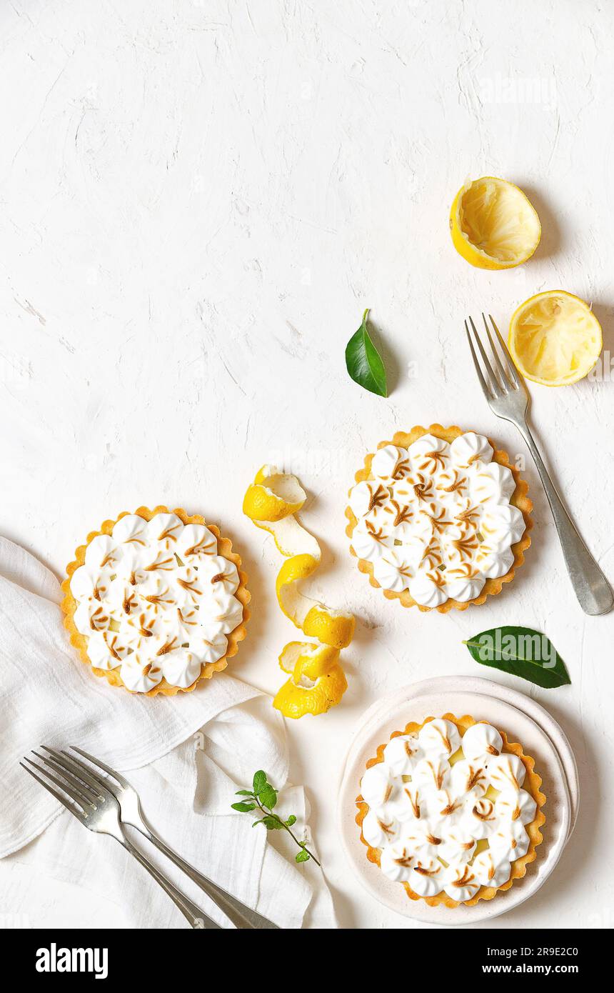 Mini-Zitronenkuchen mit Meringue, Zitronenschale, Zitronenblättern, Gabeln und einer weißen Serviette auf weißem Hintergrund mit Kopierraum. Stockfoto