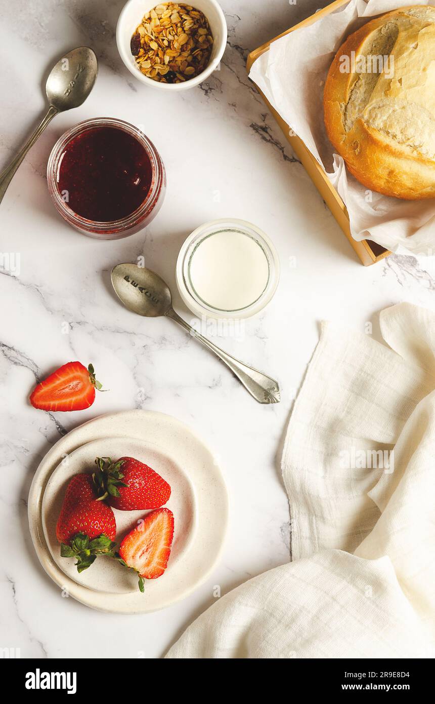 Einfacher Joghurt, frisches Obst, Erdbeermarmelade, eine Schüssel Müsli und ein Brotlaib mit einer weißen Serviette auf marmoriertem Hintergrund. Draufsicht. Stockfoto