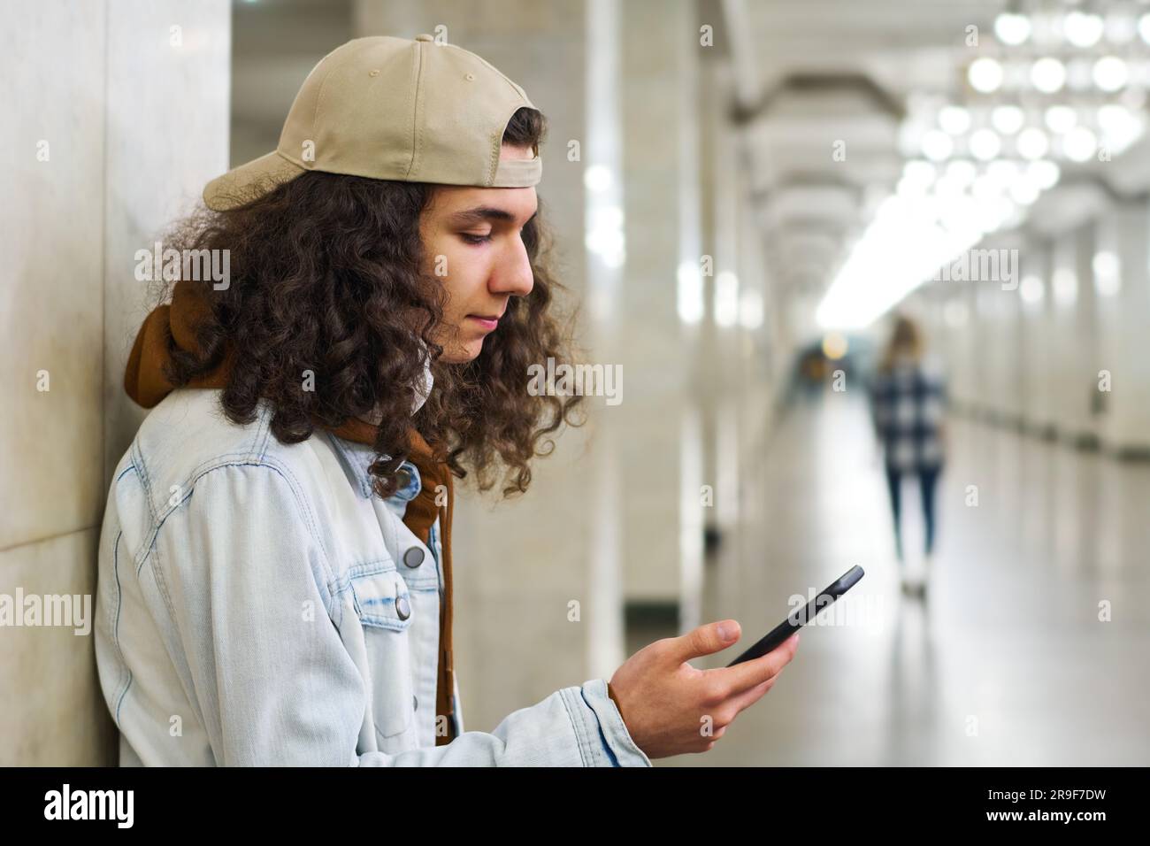Ein Teenager mit langen, dicken Locken, der SMS schreibt oder Videos auf einem Smartphone ansieht, während er auf die U-Bahn in der U-Bahn-Station wartet Stockfoto