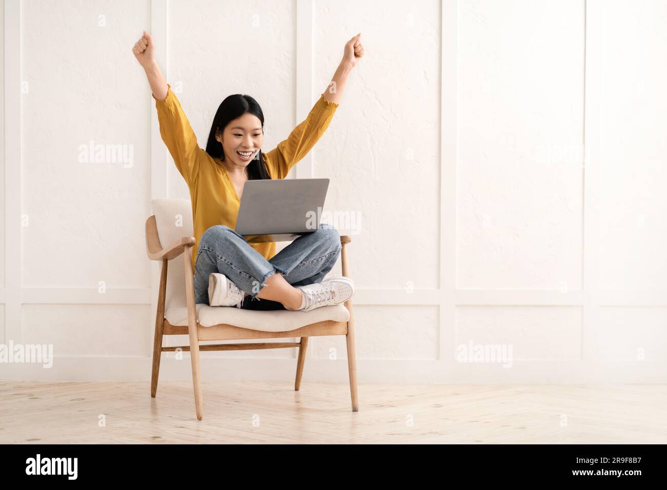 Glückliche chinesin, die im Sessel sitzt, einen Computer-Laptop benutzt, Gesten macht Stockfoto