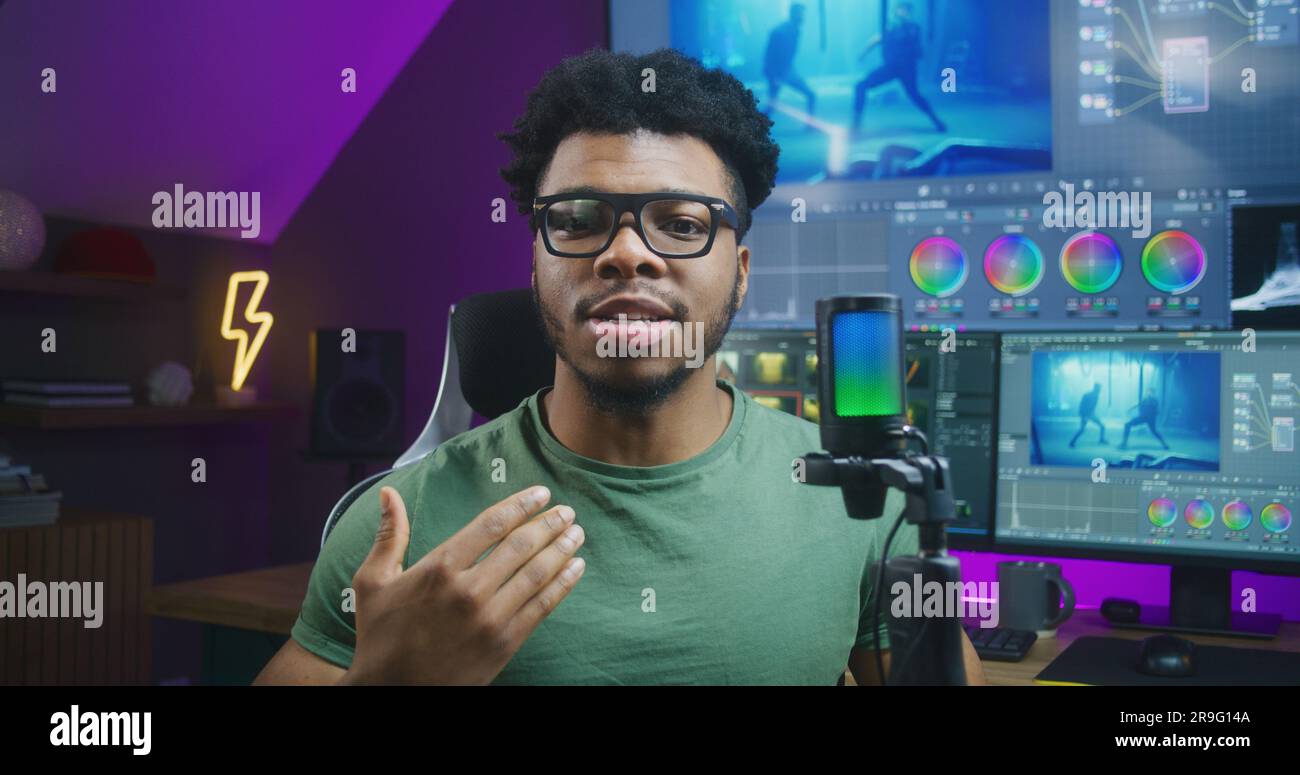 Der afroamerikanische Farbfotograf spricht mit Mikrofon vor der Kamera und nimmt Schulungsvideos zur Farbkorrektur und Videobearbeitung auf. Filmmaterial und Programmoberfläche auf Computer und Großbildschirm. Stockfoto