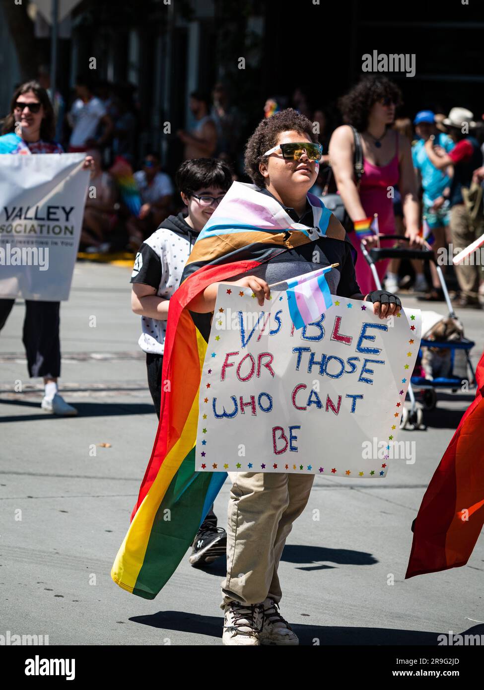 Eine junge Person mit Regenbogenflagge trägt bei der alljährlichen Sacramento-Pride-Parade eine Trans-Flagge und ein Schild mit der Aufschrift „sichtbar für diejenigen, die nicht sein können“ Stockfoto