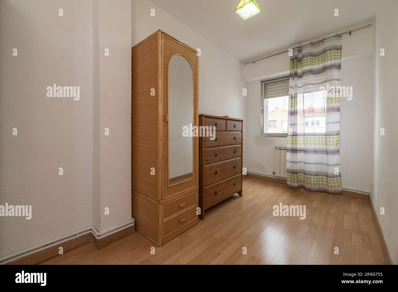 Ein teilweise leerer Raum mit einem schmalen Korbgarderobe mit Spiegel und einer Holzschränke mit Schubladen und Belüftung mit Vorhängen Stockfoto