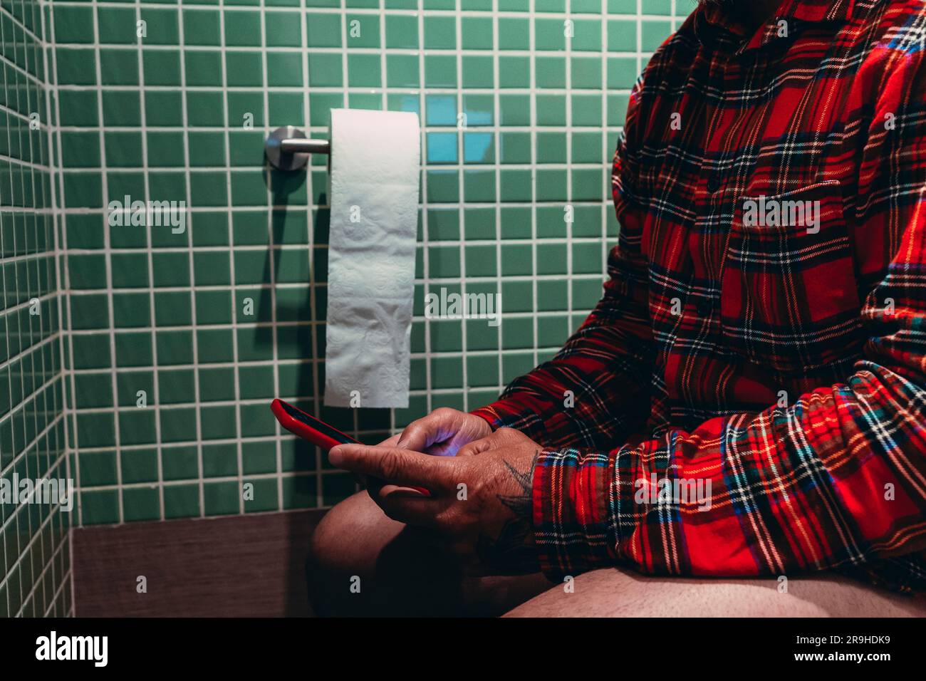 Der Mann hält sein Smartphone in der Hand, um sich die Zeit auf der Toilettenschüssel im Badezimmer zu vertreiben. Er liest die Morgennachrichten und durchsucht das Handy bef Stockfoto