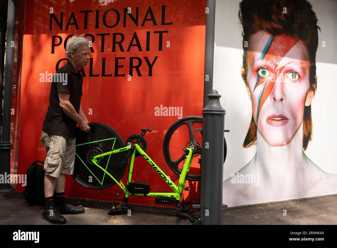 Ein Radfahrer flickt einen Reifenschaden neben dem berühmten Gesicht von David Bowie, wie er von Brian Duffy für das Cover von Bowies Aladdin Sane Album Cover fotografiert wurde, und blickt auf die Charing Cross Road im West End, am 26. Juni 2023 in London, England. Bowies Porträt ist in der nahe gelegenen National Portrait Gallery zu sehen, die kürzlich nach umfangreichen Renovierungsarbeiten wieder eröffnet wurde. Stockfoto