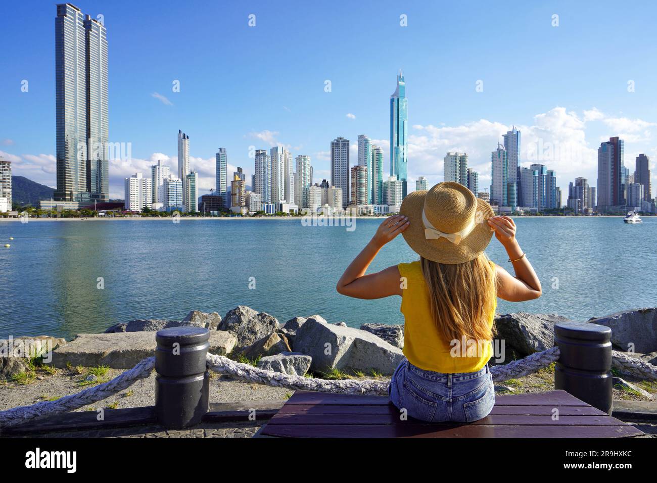 Urlaub in Balneario Camboriu, Brasilien. Rückansicht einer jungen Frau mit Hut entspannt auf einer Bank sitzen und die Skyline von Balneario Camboriu, Santa Catarina, Stockfoto