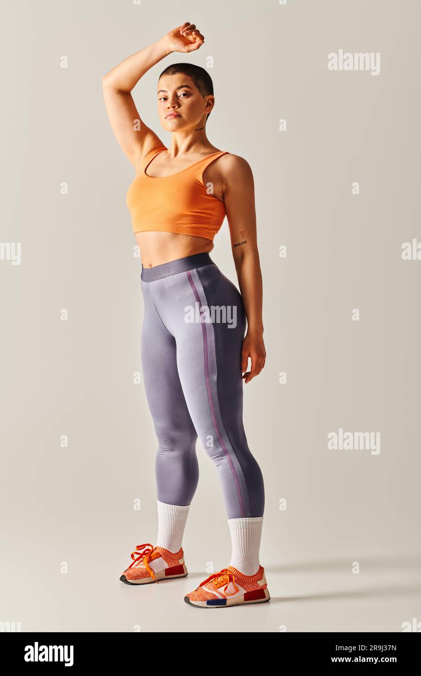 Körperbewusstsein, sportliche und kurzhaarige Frau, die auf grauem Hintergrund posiert, kurviges Fitnessmodell, mit erhobener Hand stehen, Ausdauer und Befähigung, Stockfoto