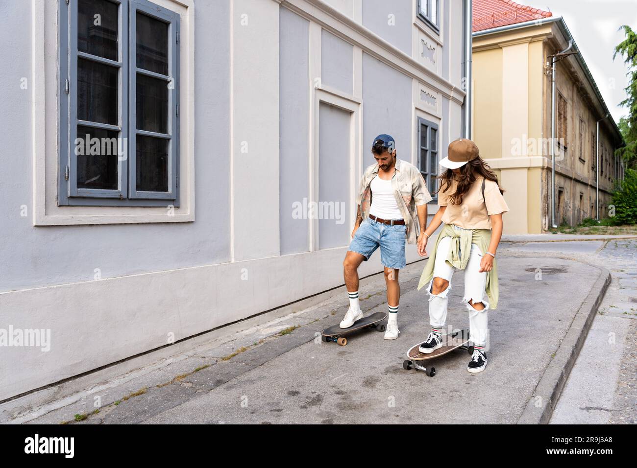 Touristen Paare Skateboarder, die Schlittschuhe fahren. Glückliche junge Reisende, die gemeinsam auf dem Asphalt bei Sonnenuntergang sitzen. Stylischer Mann und Frau in trendigen Outfits Stockfoto