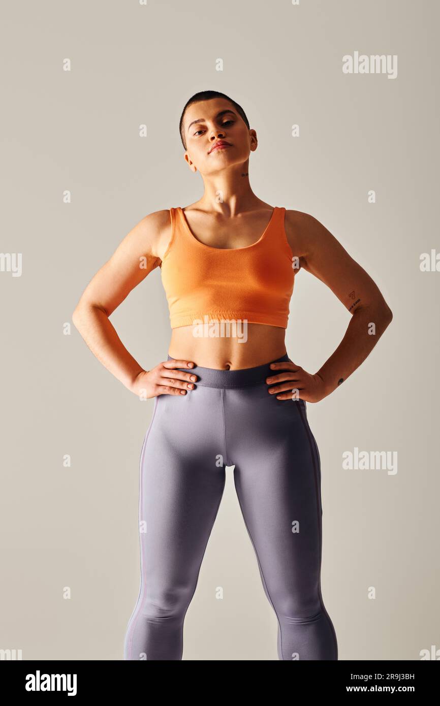 Körperbewusstsein, tätowierte und kurzhaarige Frau, die mit den Händen auf der Hüfte steht und auf grauen Hintergrund auf die Kamera schaut, kurviges Fitnessmodell im Sportprogramm Stockfoto