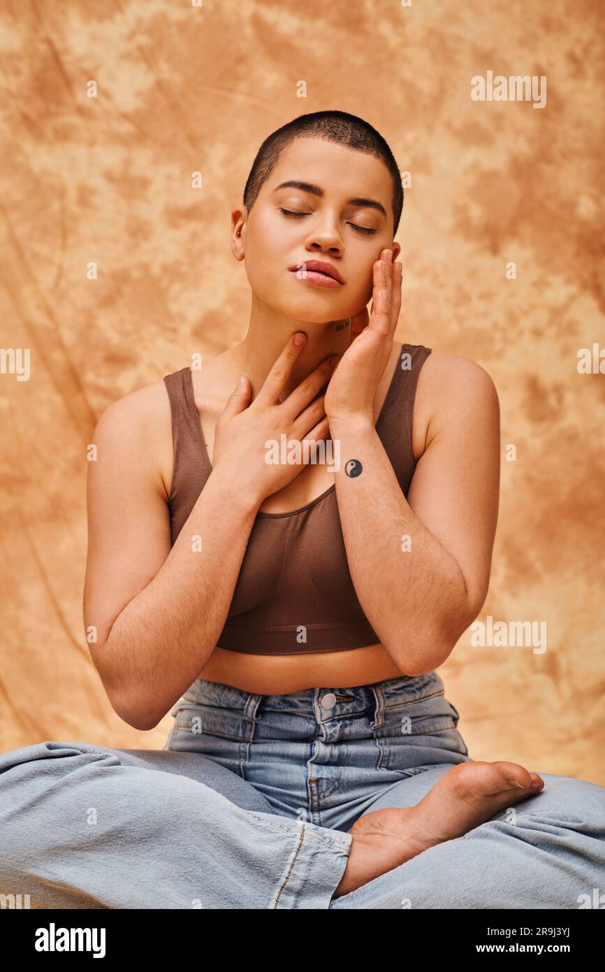 Körperbild, Entspannung, kurvige junge und tätowierte Frau in Jeans und gekreuztes Top sitzend mit gekreuzten Beinen auf meliertem beigefarbenem Hintergrund, geschlossene Augen, Persos Stockfoto