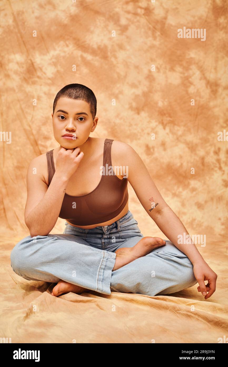 Körperbewusstsein, Akzeptanz, kurvige junge und tätowierte Frau in Jeans und Crop Top Sitting mit gekreuzten Beinen auf meliertem beigefarbenem Hintergrund, persönlicher Stil Stockfoto