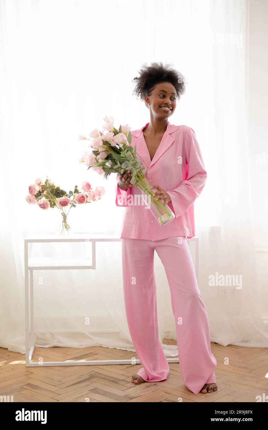 Junge afrikanische fröhliche Frau in pinkfarbenen Pyjamas, die im Zimmer mit Blumenstrauß steht und vor dem Hintergrund des Fensters lächelt. Fröhliches Ereignis, Geburtstag, holi Stockfoto