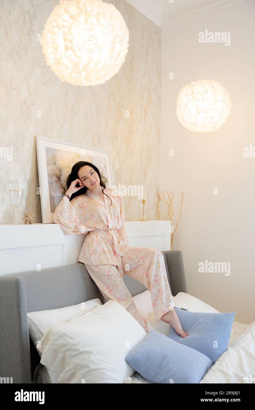 Junge, brünette Frau im Pyjama, die auf dem Bett vor dem Hintergrund von Bettwäsche und Inneneinrichtung sitzt. Stockfoto