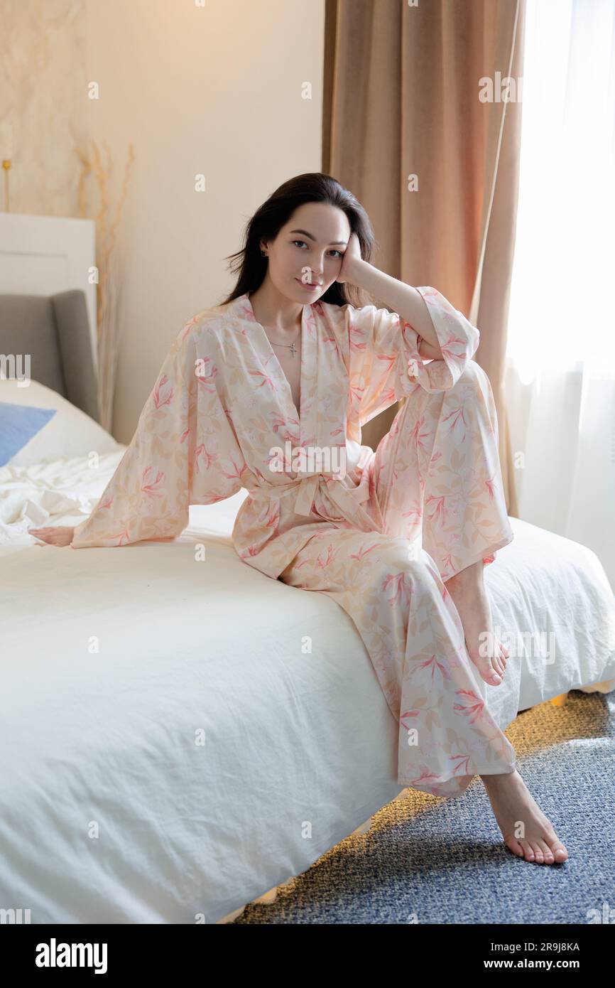 Junge brünette Frau im Pyjama, die auf dem Bett vor Bettwäsche und Fenster sitzt. Stockfoto