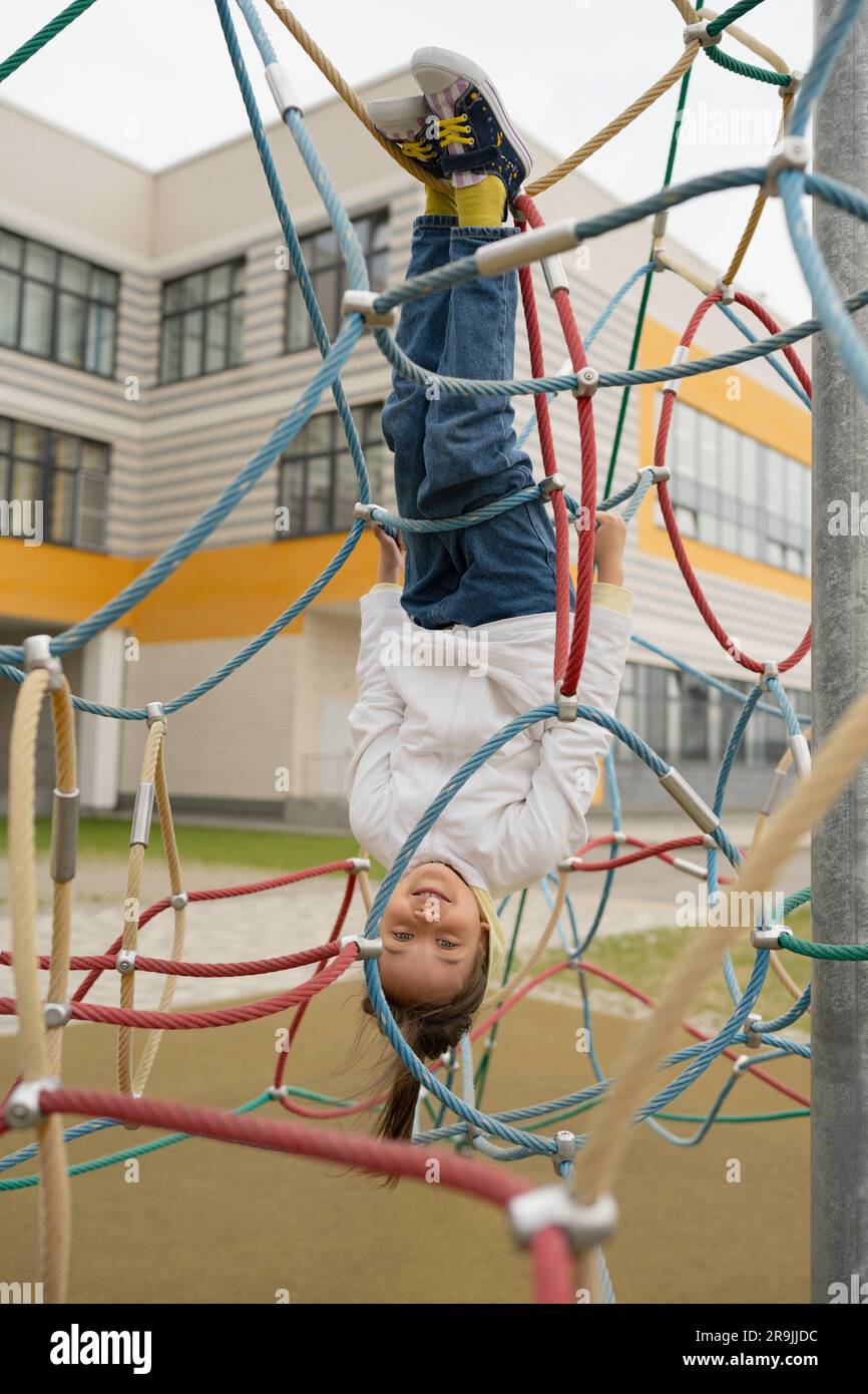 Ganzkörperansicht eines positiven Mädchens in lässiger, bequemer Kleidung, die kopfüber an Seilen hängt, während du draußen auf dem Sportplatz spielst Stockfoto