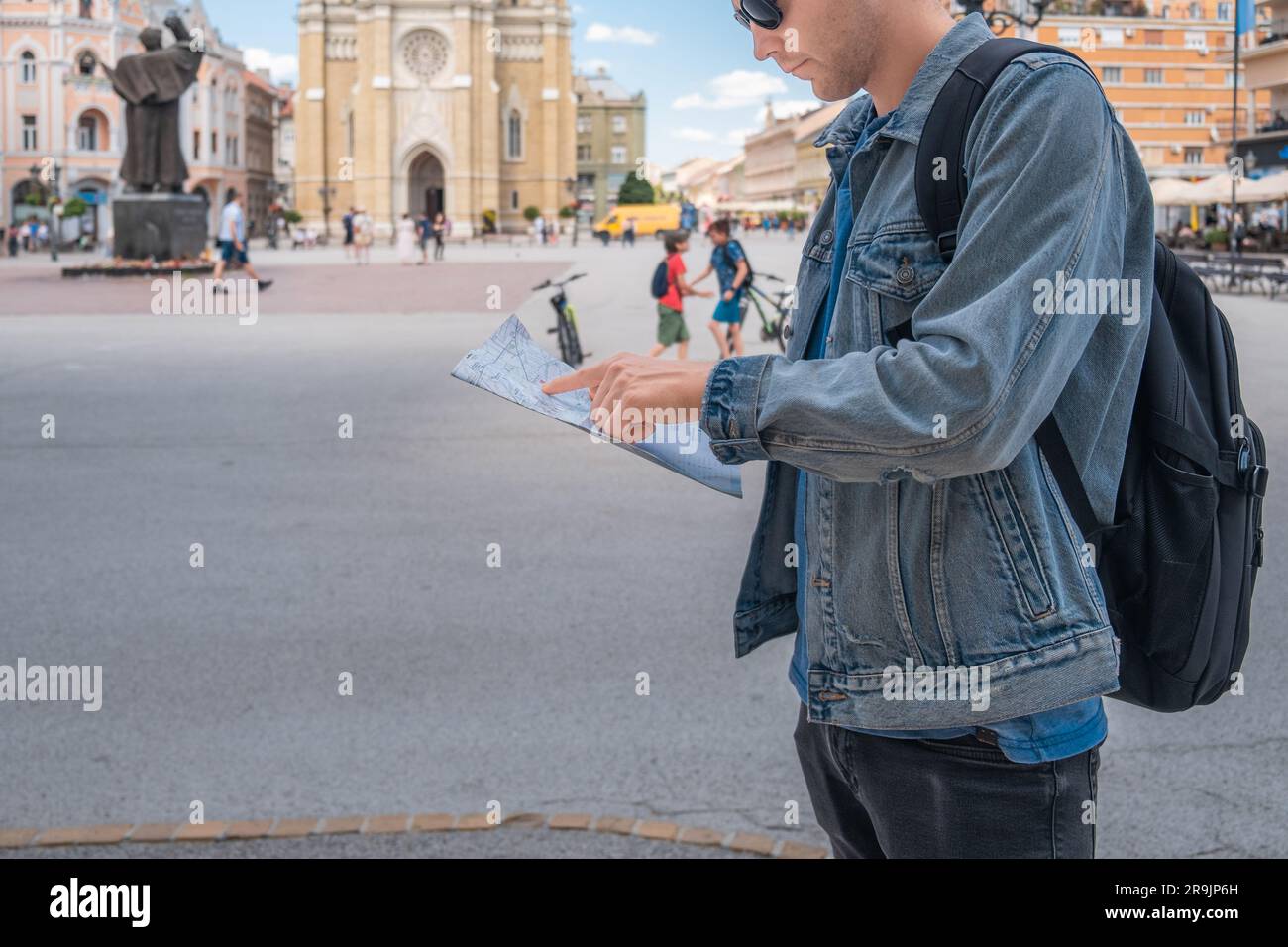 Ein Tourist auf dem Platz in der Stadt, der auf einer Karte aus Papier steht, erforscht die neue Stadt. Hübscher männlicher Reisender Rucksacktourist mit Sonnenbrille Stockfoto