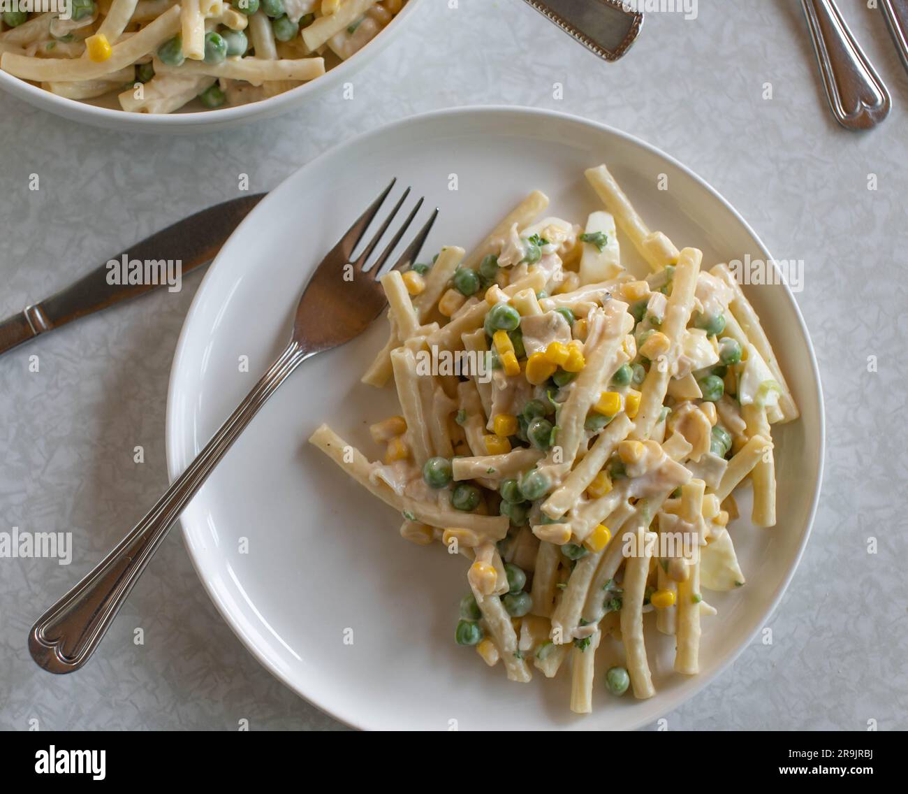 Teller mit klassischem Nudelsalat. Mit Mayonnaise, gekochten Eiern, grünen Erbsen, Mais und Wurst auf einem Tisch Stockfoto