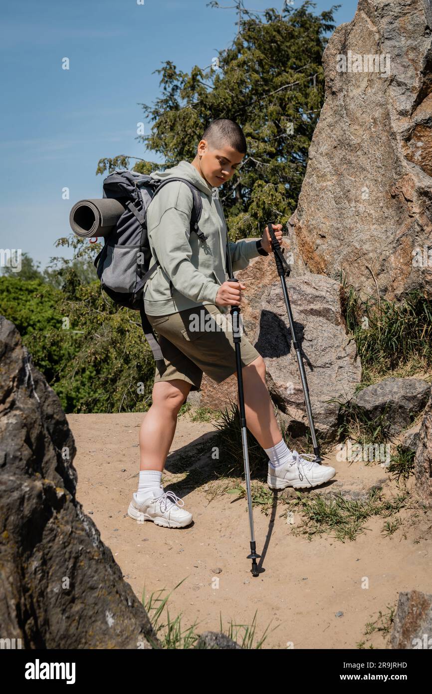 Junge, kurzhaarige Wanderer mit Rucksack, der Wanderstöcke hält und den Weg betrachtet, während sie zwischen Steinen mit blauem Himmel im Hintergrund stehen, tr Stockfoto