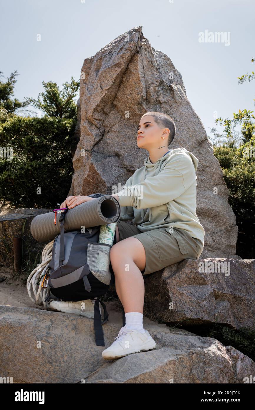 Junge Frau mit kurzen Haaren in lässiger Kleidung, die neben dem Rucksack sitzt, mit Karte auf Steinen auf einem Hügel mit Natur im Hintergrund, ruhige Wanderer, die innere sportarten finden Stockfoto