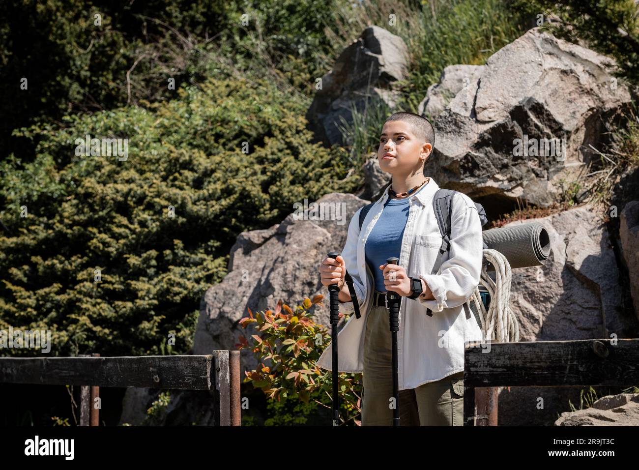 Junge, kurzhaarige Touristin mit Rucksack und Kletterseil, die Wanderstöcke hält, während sie wegschaut und mit der Natur im Hintergrund steht, wo Stockfoto