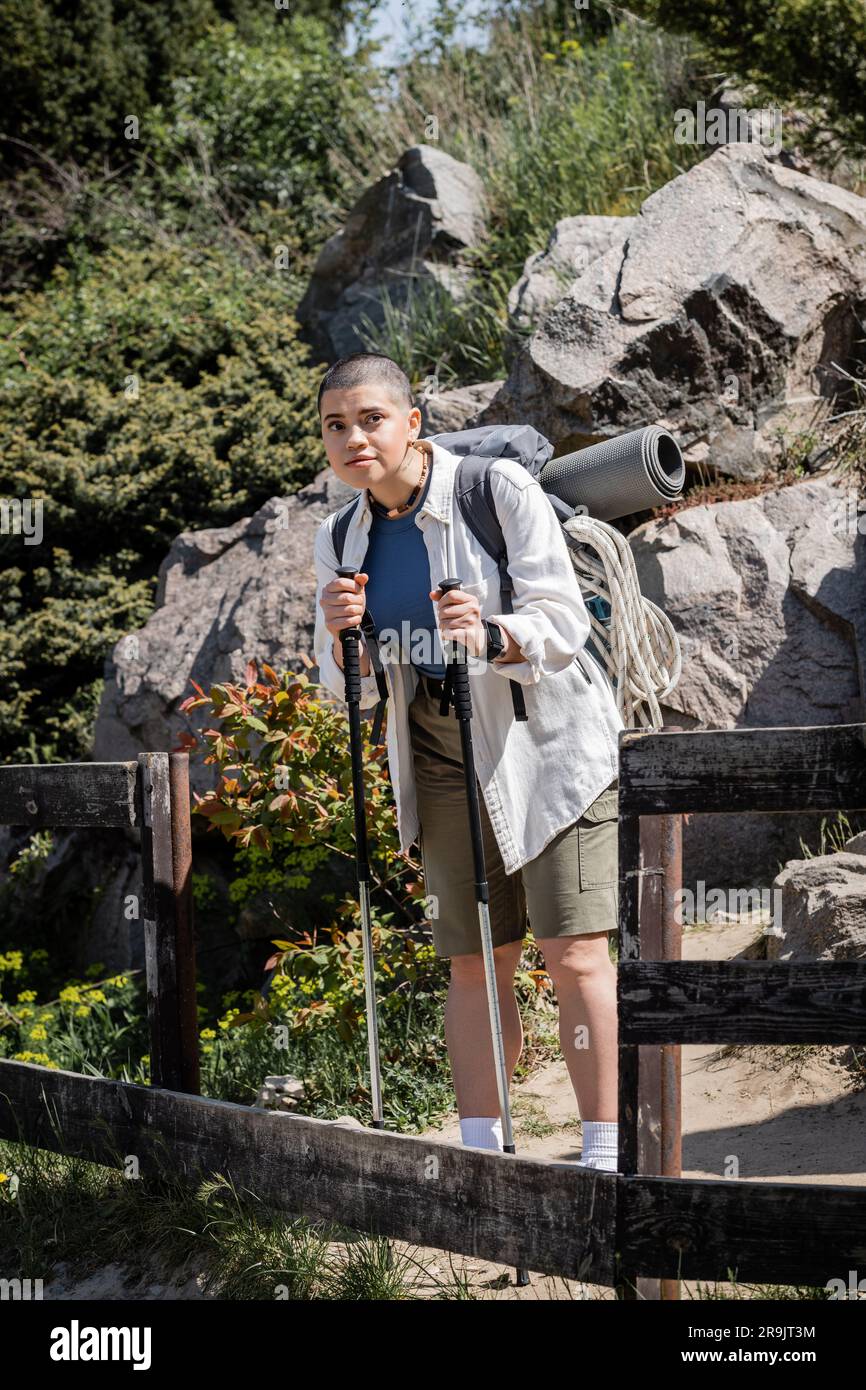 Junge, kurzhaarige Frau, die in lässiger Kleidung unterwegs ist, mit Rucksack, der Wanderstöcke hält und wegschaut, während sie mit der Natur im Hintergrund steht, wo Stockfoto