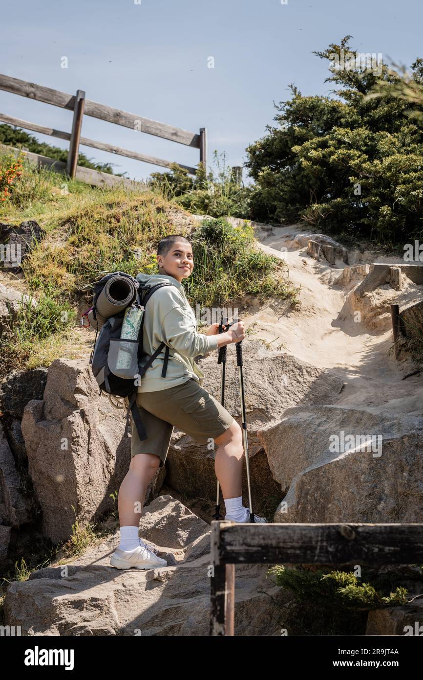 Positive junge, kurzhaarige Frau mit Rucksack, der Wanderstöcke hält und im Sommer in der Nähe des Hügels mit Steinen im Hintergrund steht, Trans Stockfoto