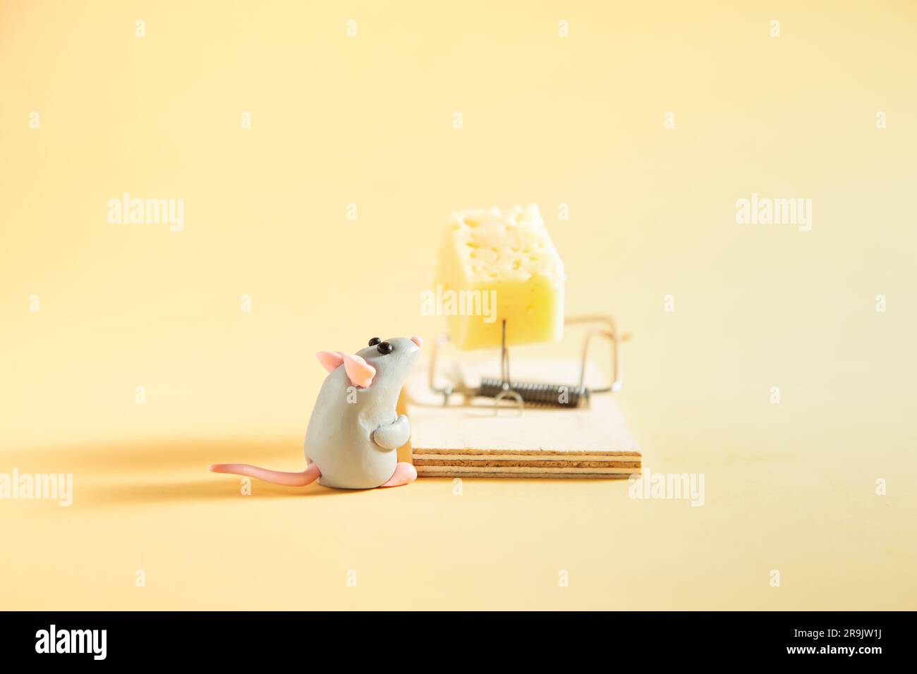 Eine kleine, graue Plastikmaus sieht sich ein Stück Käse in einer Mausefalle an. Beiger Hintergrund. Stockfoto