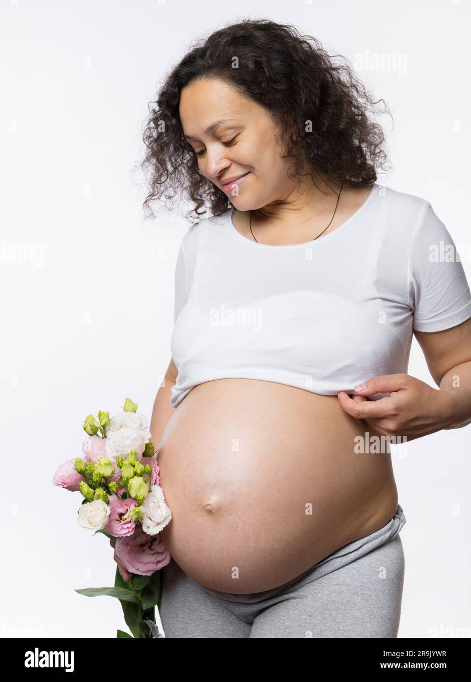 Wunderschöne, multiethnische, gravide Frau, die einen Blumenstrauß in der Hand hält, lächelt und ihren schwangeren Bauch in der späten Schwangerschaft ansieht Stockfoto