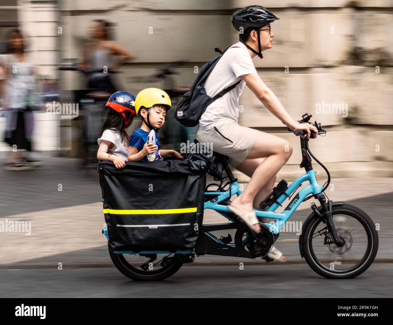 Ein Mann, der mit zwei Kindern auf einem Cargo-Bike-Anhänger hinten fährt, vorbei an V&A in London, verschwommen durch Geschwindigkeit Stockfoto