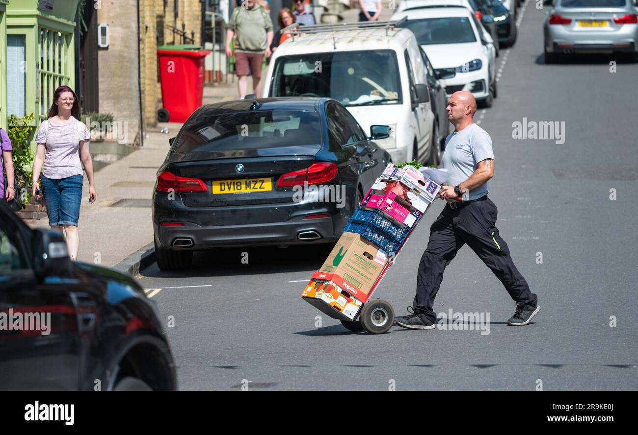 Lieferfahrer schiebt Transportwagen über eine Straße und liefert Lebensmittel an einen Einzelhändler in England. Stockfoto