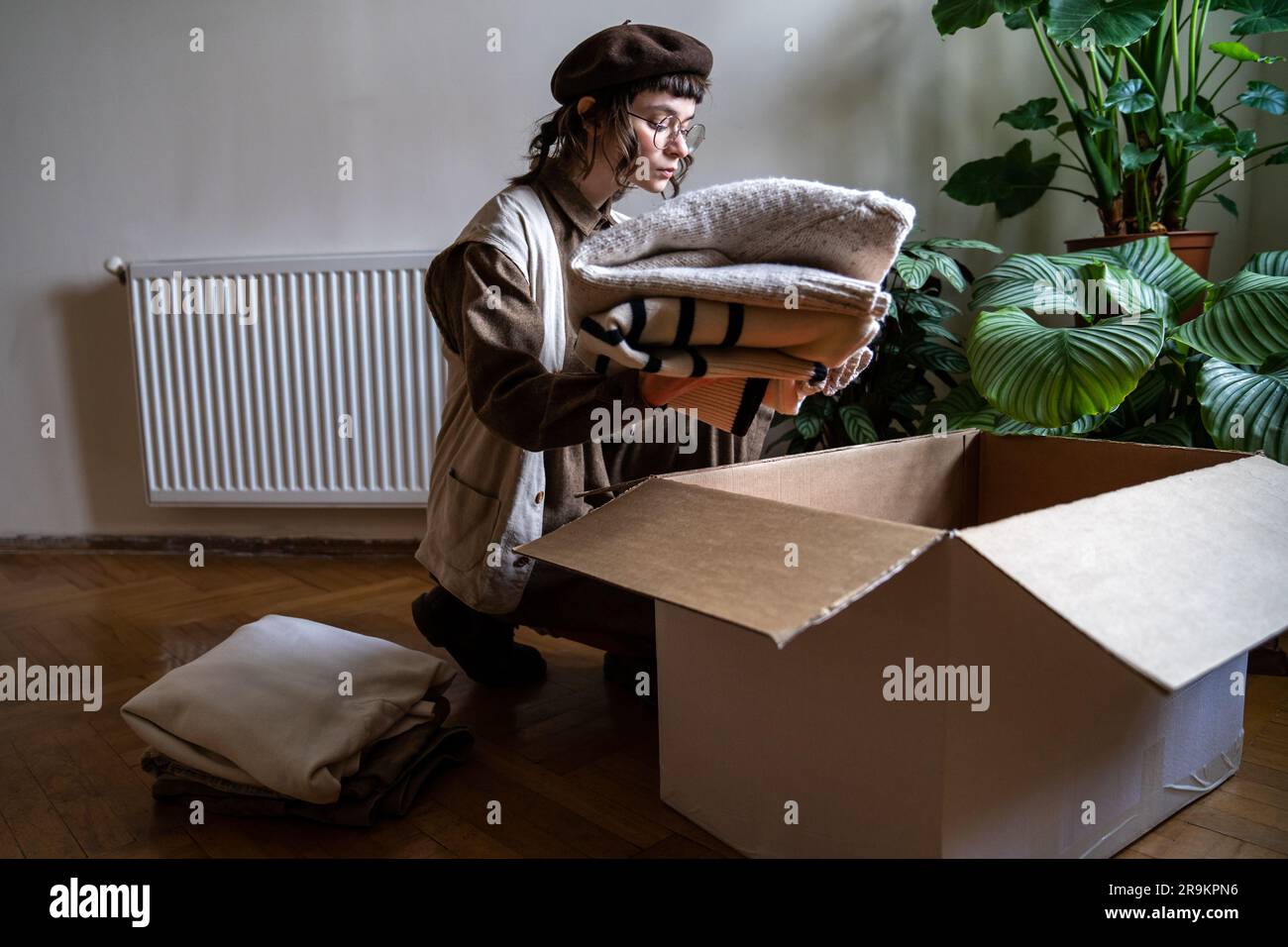 Caring volunteer girl in eyeglasses puts used clothes in cardboard box. Conscious consumption things Stockfoto