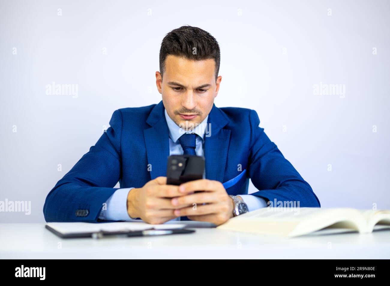 Attraktiver Geschäftsmann, Wirtschaftswissenschaftler, Anwalt, Manager oder Geschäftsführer, der mit einem Smartphone tippt und Finanzdokumente am Schreibtisch prüft. Stockfoto