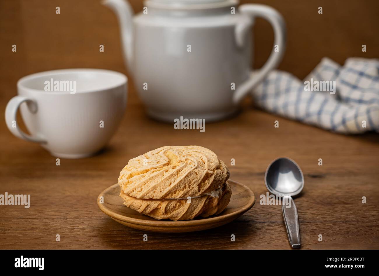 Szene der Teezeit mit einem kleinen, hausgemachten runden Kuchen gefüllt mit Blaubeercreme in einem Holzgericht mit einer weißen Keramiktasse Tee und Metalllöffel Stockfoto