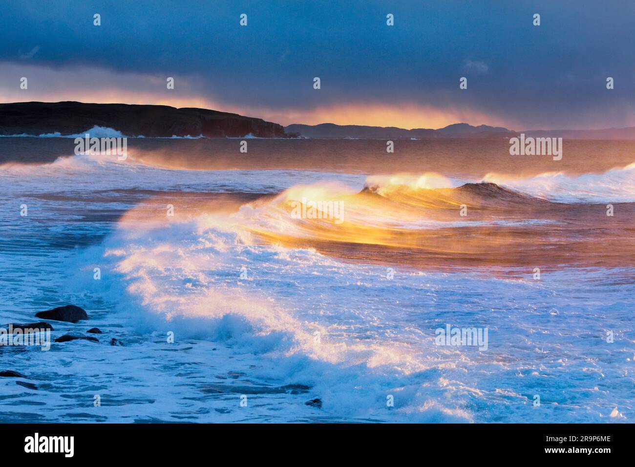 Große Wellen, die während eines Wintersturms stürzen, das wirbelnde Spray erhellt durch das warme Licht der Abendsonne. Im Hintergrund Summer Isles, nahe Reiff an der Nordwestküste Schottlands. Stockfoto