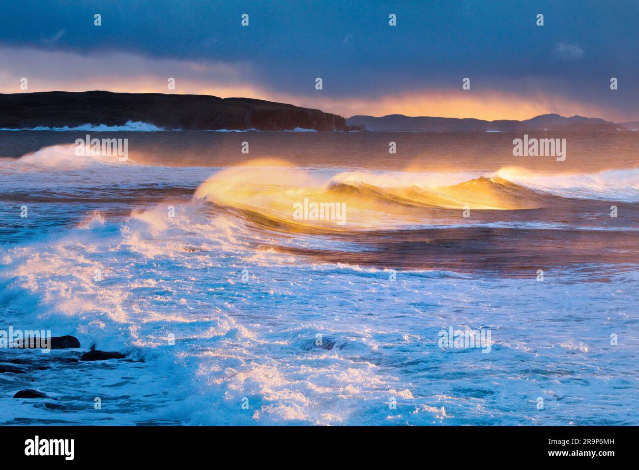 Große Wellen, die während eines Wintersturms stürzen, das wirbelnde Spray erhellt durch das warme Licht der Abendsonne. Im Hintergrund Summer Isles, nahe Reiff an der Nordwestküste Schottlands. Stockfoto