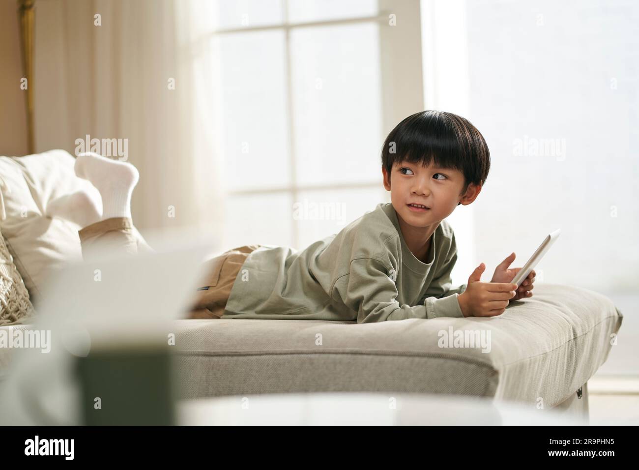 Süßer kleiner asiatischer Junge, der zu Hause vorne auf dem Familientisch liegt und ein digitales Tablet hält Stockfoto