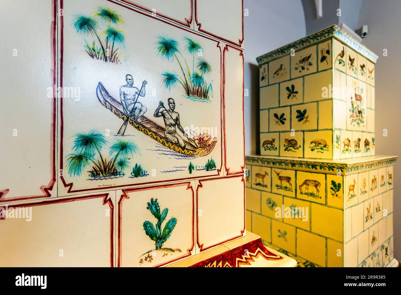 Ein handbemalter Kachelofen im Porzellanmuseum der Porzellan-Stiftung Meissen, Sachsen, Deutschland, zeigt zwei Männer in einem Kanu zwischen Palmen und Kaktusmotiven auf einer Kachel. Daneben steht ein zweiter, gelb glänzender Kachelofen, der mit Tier- und Blattmotiven verziert ist. Stockfoto