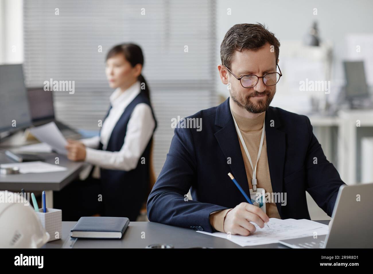 Porträt eines bärtigen männlichen Ingenieurs, der Computer mit Bauplänen und Plänen benutzt, während er Berechnungen im Büro und im Kopierraum macht Stockfoto