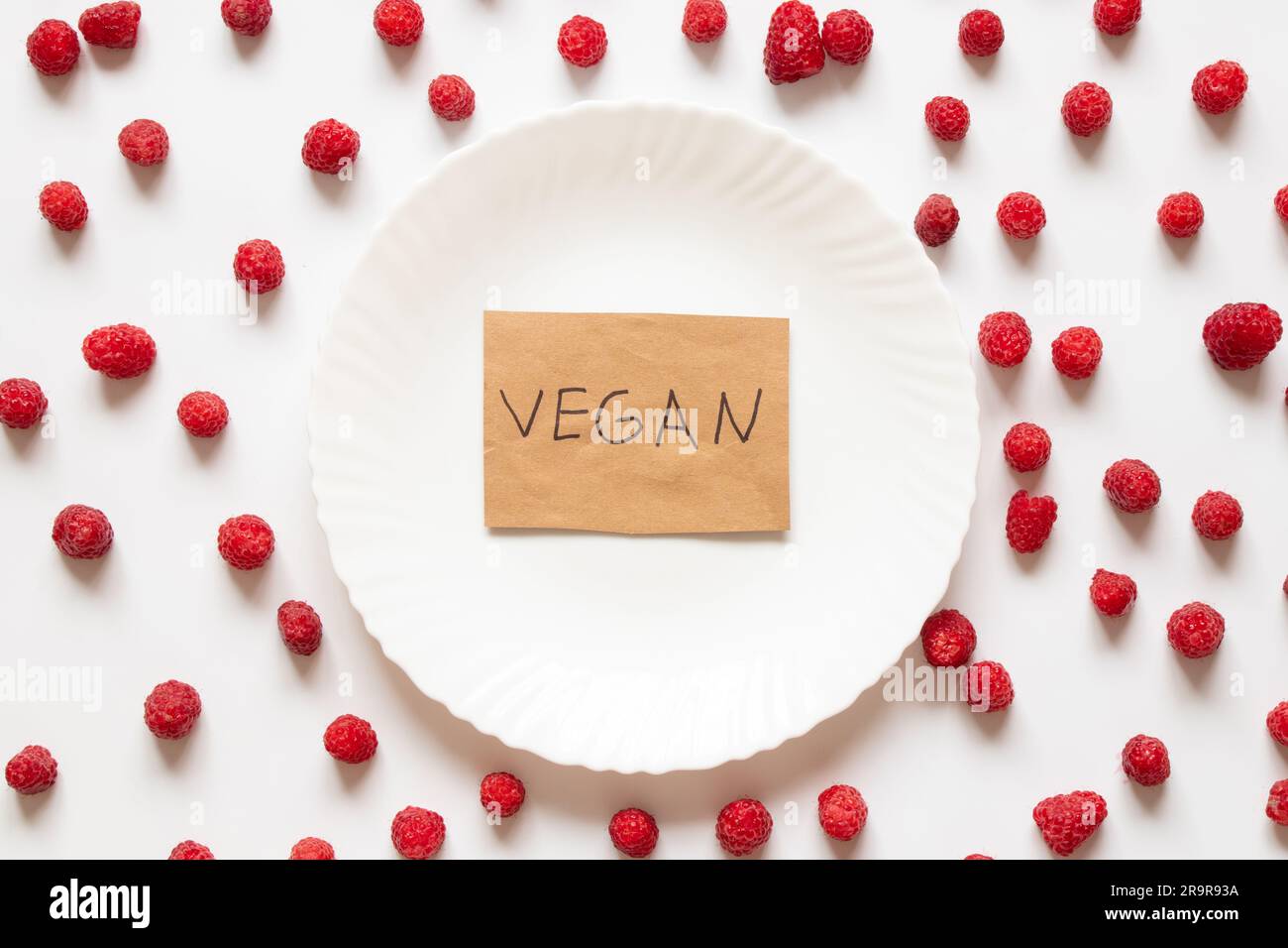 Das Wort vegan ist auf Papier geschrieben und liegt auf einem Teller mit Himbeeren auf weißem Hintergrund, ohne Fleisch und Früchte Stockfoto