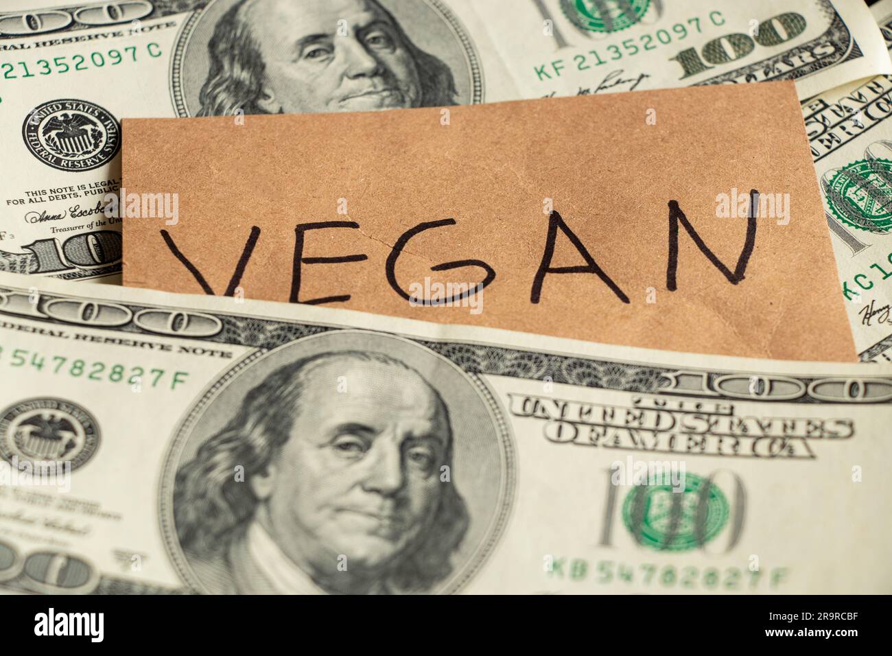 Das Wort vegan steht auf dem Papier, auf dem Hintergrund eines 100-Dollar-Scheins, der Ablehnung von Fleisch, dem Preis von Gemüse, dem Geschäft Stockfoto