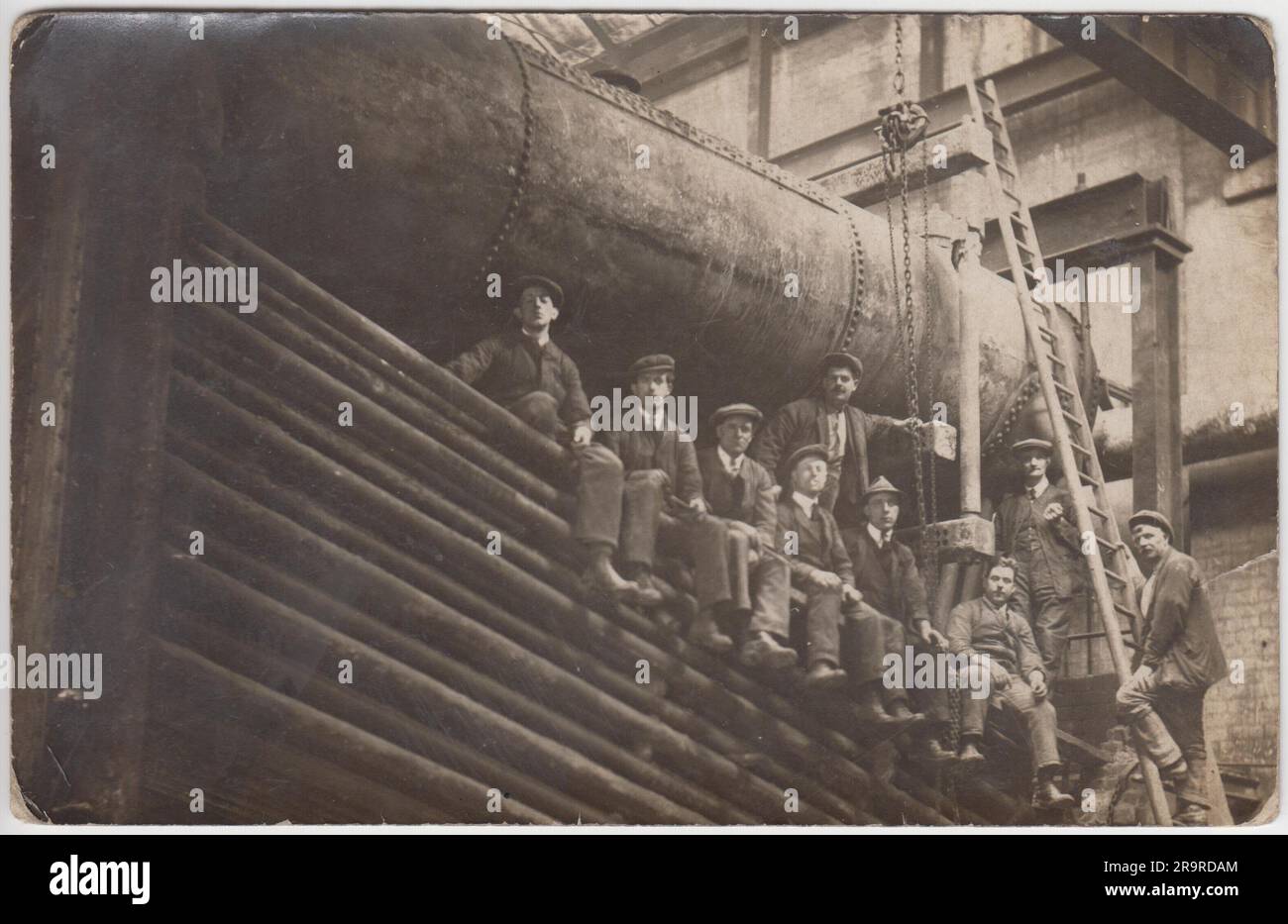 Enginemen: Foto einer Gruppe von Männern aus dem frühen 20. Jahrhundert, die neben einer großen Turbine oder einem Motor in einer Fabrik auf Rohren sitzen und stehen Stockfoto