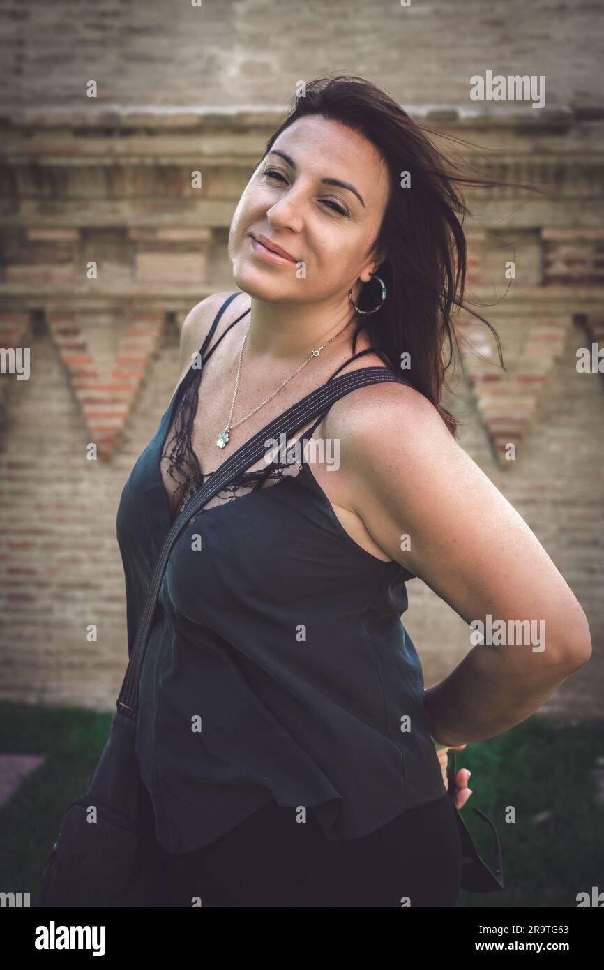 Halbkörperporträt einer lächelnden brünetten Frau im Freien, mit dem Hintergrund einer Mauer aus Ziegelstein, die Authentizität und Freude vermittelt. Stockfoto