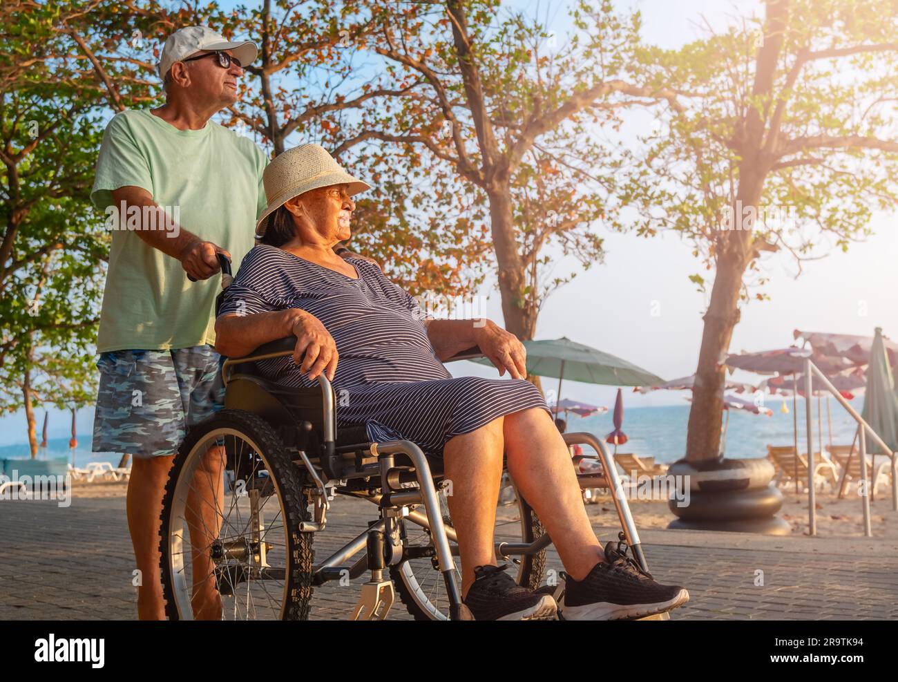 Ein Senior trägt seine Frau im Rollstuhl für einen Spaziergang im Freien Stockfoto