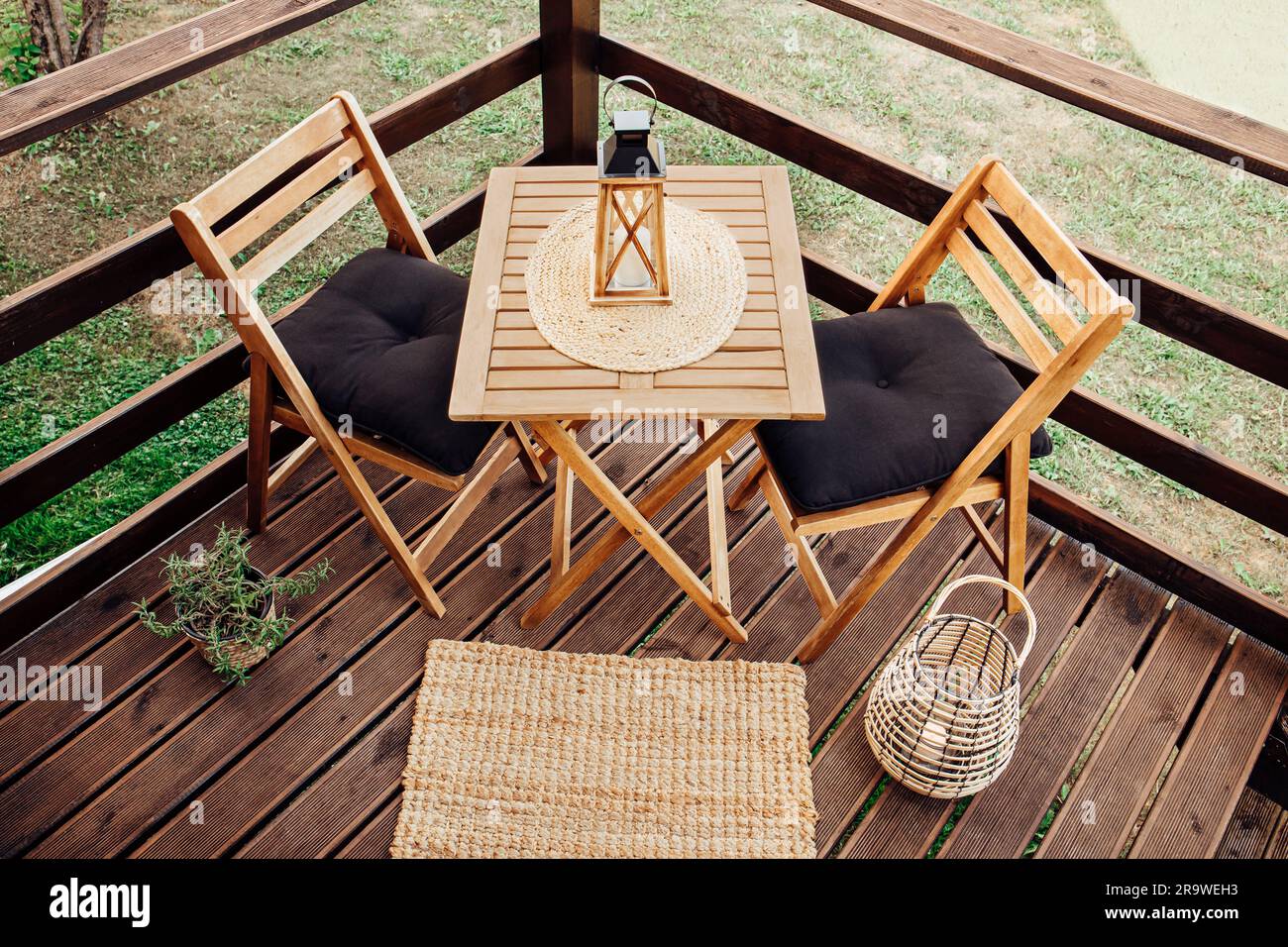 Moderner, kleiner Holzbalkon mit Sitzecke, Gartenmöbeln aus Holz, weichen schwarzen Kissen, Laterne mit brennender Kerze auf dem Tisch im Freien. Stockfoto