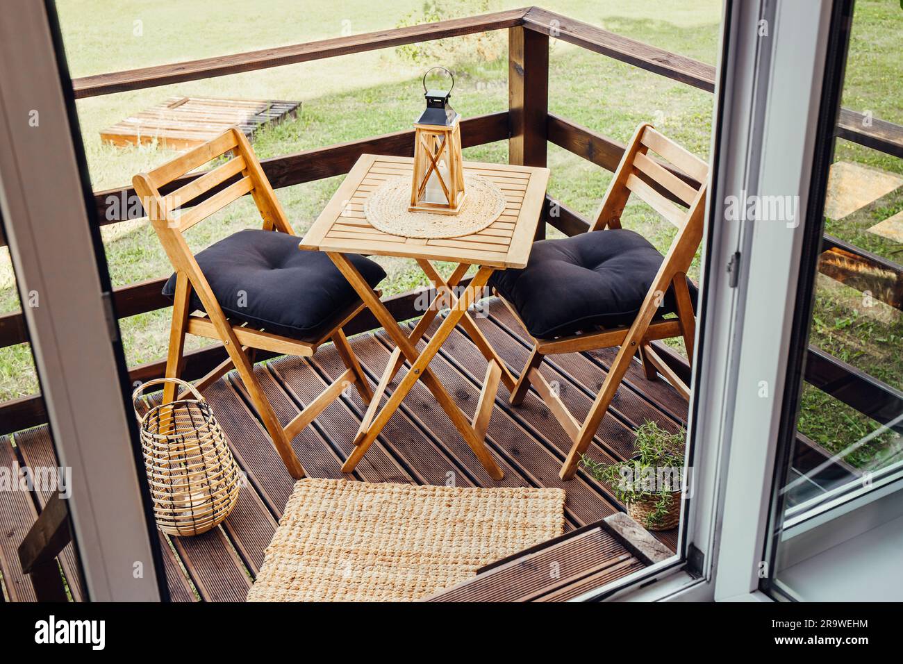 Moderner, kleiner Holzbalkon mit Sitzecke, Gartenmöbeln aus Holz, weichen schwarzen Kissen, Laterne mit brennender Kerze auf dem Tisch im Freien. Stockfoto