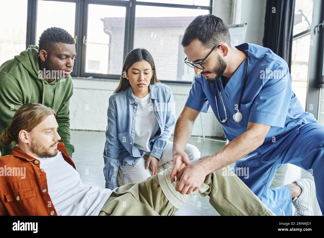 Professionelles Gesundheitspersonal, das Kompressionsverband auf das Bein des Mannes in der Nähe von multiethnischen Schülern während des erste-Hilfe-Seminars im Trainingsraum, importa anlegt Stockfoto