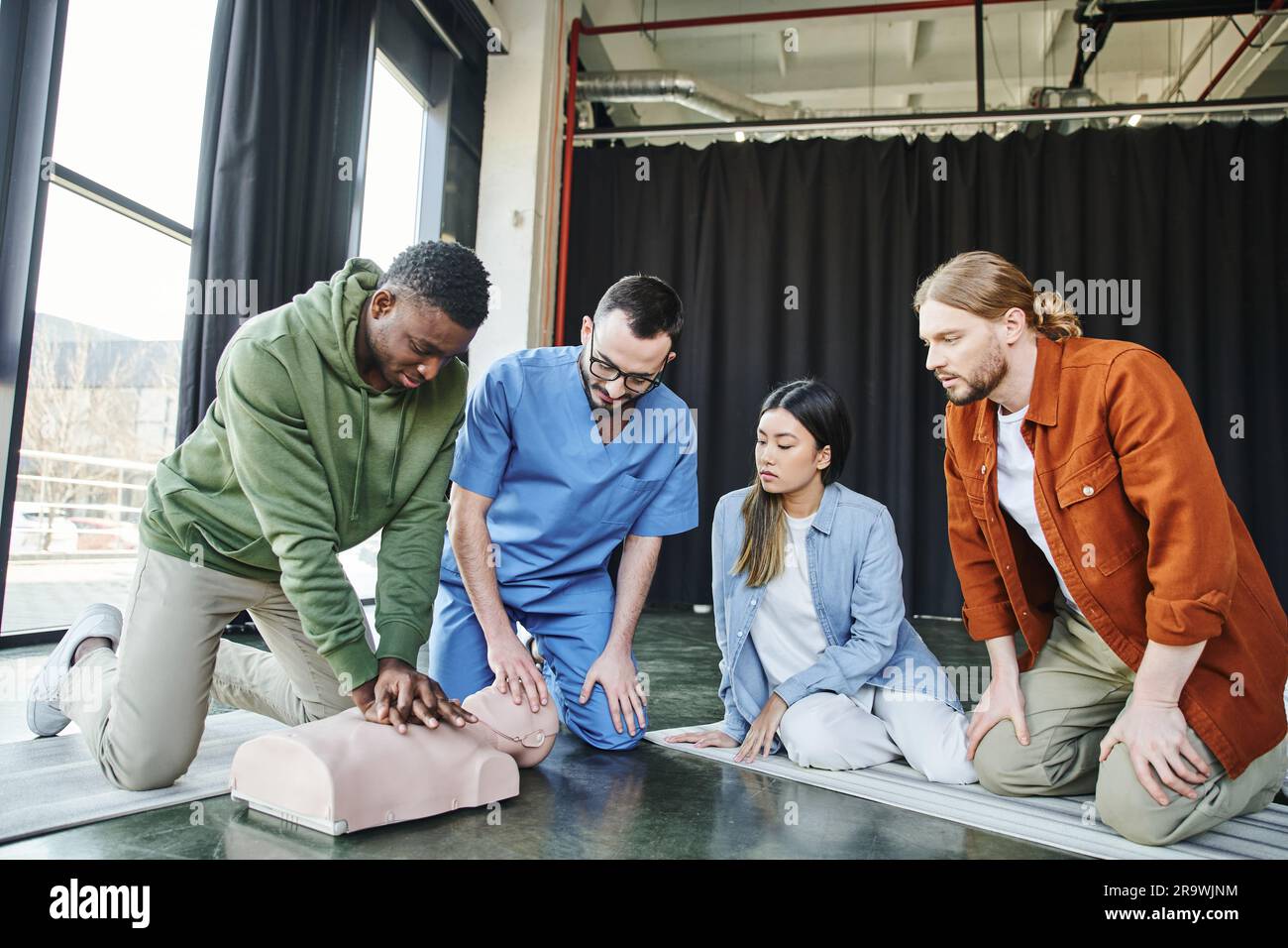 ein afroamerikanischer Mann führt Thoraxkompressionen an der HLW-Prüfpuppe durch, während er beim ersten-Hilfe-Seminar in der Nähe eines professionellen Sanitäters und einer vielfältigen Gro lernt Stockfoto