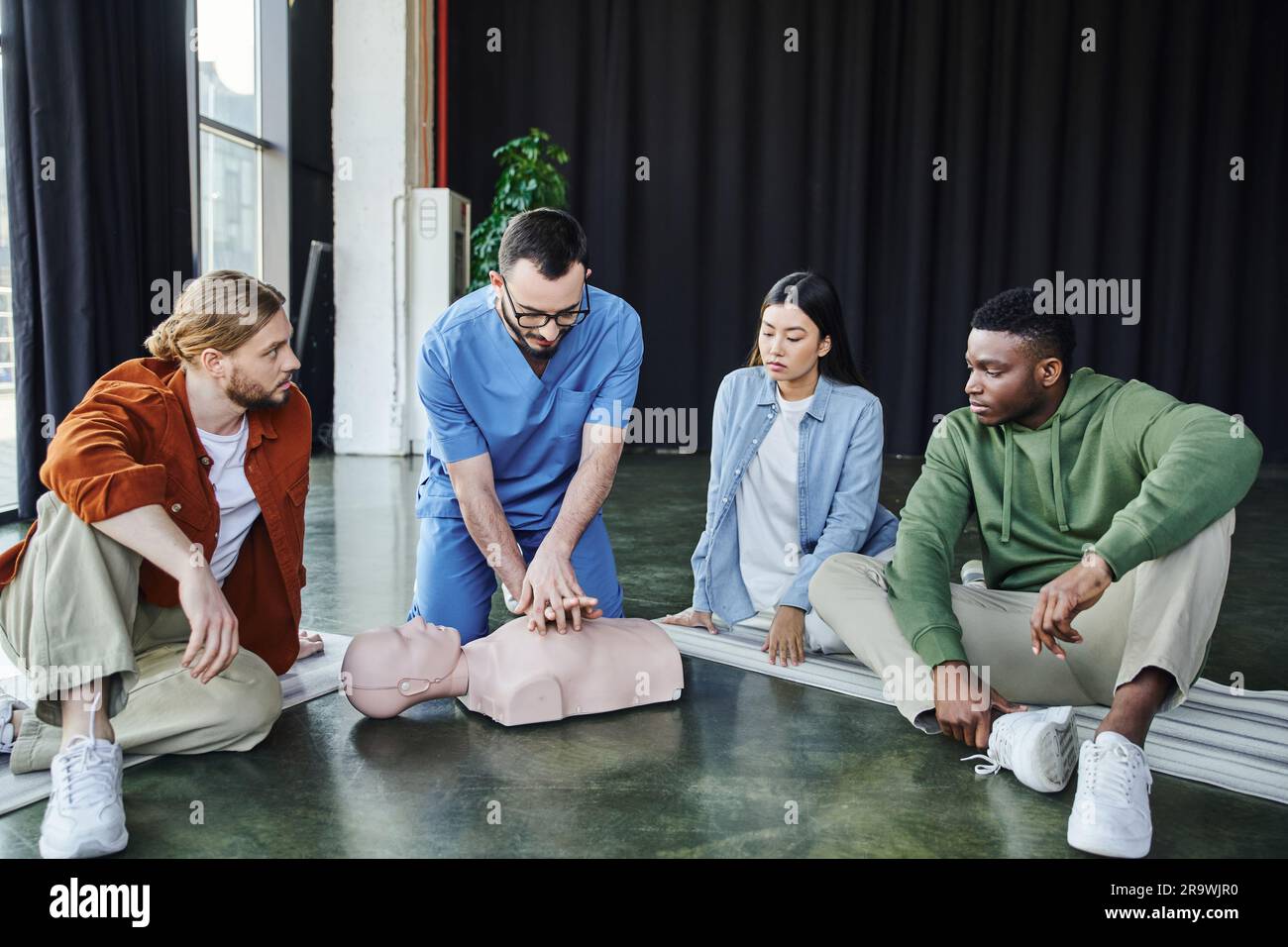 Professioneller Sanitäter, der Thoraxkompressionen an der HLW-Prüfpuppe durchführt, während er dem jungen multikulturellen Team Dura kardiopulmonale Reanimationstechniken zeigt Stockfoto