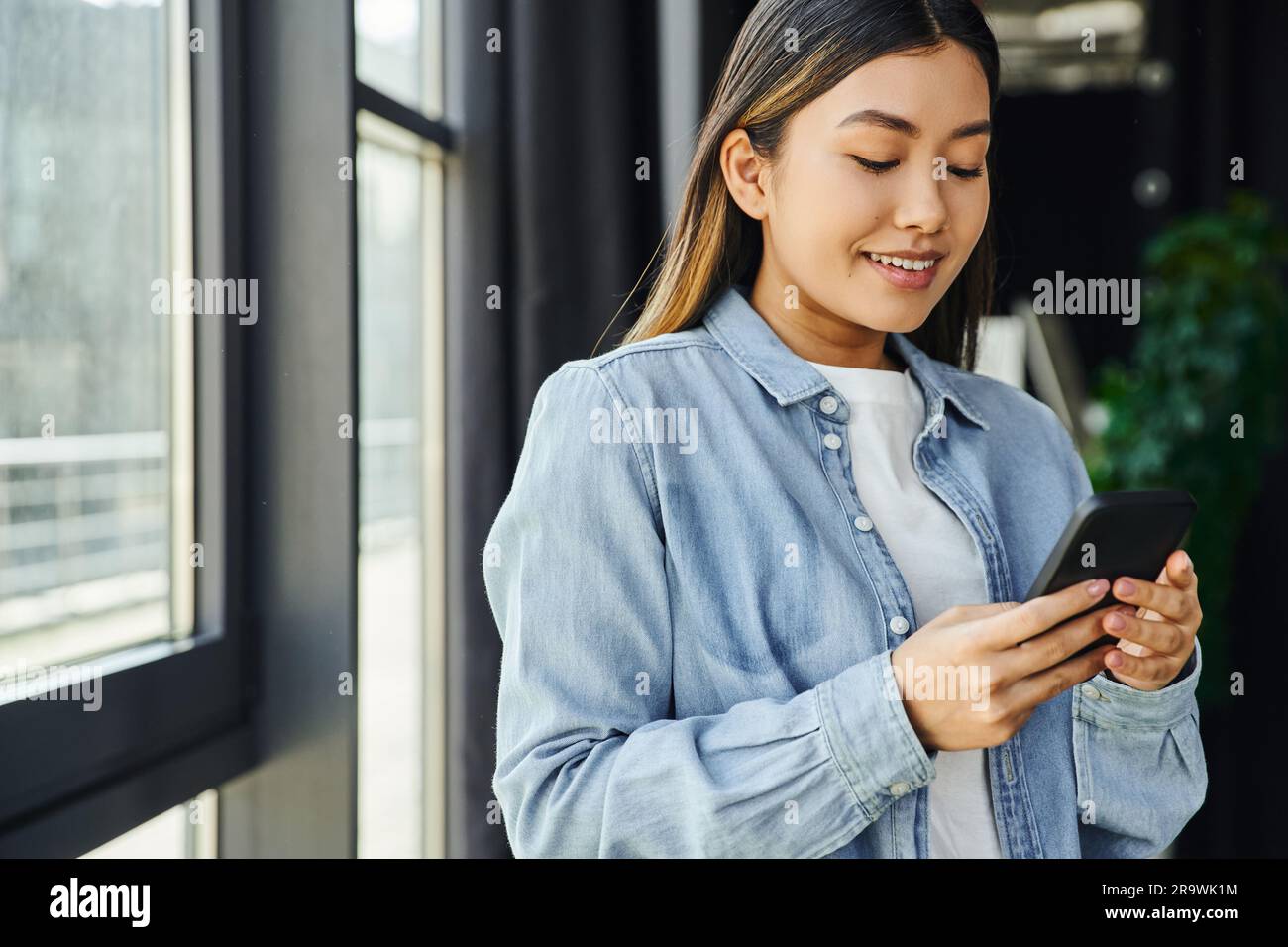 Glückliche und trendige asiatische Frau in blauem Jeansshirt, mit braunem Haar, das in einem modernen Büro steht, lächelt und Nachrichten auf dem Handy sendet, erfolgreich Stockfoto